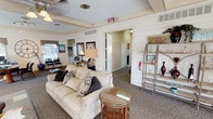 This single-floor space offers a warm and inviting atmosphere enhanced by thoughtful design details throughout. The interior is characterized by white shiplap-style wall paneling and beamed ceilings that lend a classic, cottage-inspired aesthetic across the entire layout.

The centerpiece of the main living area is a commanding floor-to-ceiling stone fireplace, featuring a broad slate hearth and a white wood mantle accented by wall sconces and framed artwork above. Comfortable seating, including a plush neutral sofa with decorative pillows and a wingback armchair, is arranged to take full advantage of this focal point. A glass-topped coffee table anchors the seating arrangement, while an open metal shelving unit displays decorative accessories throughout the reception area.

Adjacent to the main lounge, a functional workspace is equipped with a desk, office seating, and a large decorative wall clock, creating a productive yet aesthetically cohesive area. Ceiling fans provide comfortable airflow throughout.

A dining area features a round glass-top table surrounded by dark wood chairs, positioned beside a sliding glass door that floods the space with natural light and provides direct access to the exterior. Patterned carpeting runs throughout, tying the spaces together cohesively. Additional rooms are accessible via interior doorways, rounding out this well-appointed and versatile space.