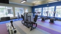 This fitness center offers a thoughtfully designed workout environment spread across a single floor. The space is anchored by a colorful striped flooring in neutral, blue, and purple tones, adding a dynamic visual element while defining different workout zones. Large windows line multiple walls, flooding the interior with natural light and providing open views of the surrounding outdoor area, creating an airy and motivating atmosphere.

The ceiling features sleek, modern pendant lighting fixtures that complement the clean architectural lines of the space. A bold navy blue accent wall adds a striking contrast to the light neutral tones of the remaining walls, contributing to an energetic yet focused ambiance.

The fitness center is outfitted with a comprehensive selection of commercial-grade equipment, including strength training machines such as a leg press and cable machines, as well as cardio equipment including treadmills, an elliptical, and a stair climber. The layout is spacious and well-organized, allowing for comfortable movement between machines. Overall, the space strikes a balance between functionality and modern design, making it a practical and visually appealing fitness facility.
