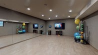 This single-floor fitness studio is thoughtfully designed to support a wide range of workout activities in a clean, modern environment. The space features light hardwood-style flooring throughout, providing a durable yet attractive surface suited for both cardio and group exercise sessions. Neutral taupe-toned walls and recessed ceiling lighting create a focused, energizing atmosphere.

The studio is divided into two functional zones. One area is dedicated to cycling, housing a row of professional-grade spin bikes positioned on protective floor mats. A wall-mounted flat-screen TV accompanies this section, allowing riders to follow along with guided workouts. A pass-through window and glass-paneled door provide connectivity to adjacent areas.

The adjacent open-floor exercise area is expansive, offering ample space for group fitness classes or individual training. A large flat-screen TV is mounted prominently on the wall, supplemented by additional smaller displays, enabling on-demand fitness programming. Full-length mirrors line one wall, enhancing the sense of space and allowing users to monitor their form. Storage cubbies hold an organized selection of dumbbells and resistance equipment, while colorful stability balls of varying sizes are neatly racked in the corner. The combination of practical equipment, smart layout, and modern finishes makes this studio a comprehensive and inviting fitness facility.