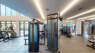 This contemporary fitness center is thoughtfully designed across a single floor, offering a comprehensive range of workout zones to accommodate diverse training needs. The facility is divided into several distinct areas, each purpose-built to maximize functionality and user experience.

The cardio zone features a generous row of treadmills and elliptical trainers positioned alongside floor-to-ceiling windows, flooding the space with natural light and offering outdoor views — an energizing environment for endurance training. Soaring double-height ceilings in this section are highlighted by striking geometric LED light fixtures that lend a modern, architectural character to the room.

The strength training area is outfitted with premium Matrix cable and functional trainer machines, free weights including a full dumbbell rack, adjustable benches, and multi-functional power racks. Recessed linear LED lighting runs throughout, maintaining a clean, polished aesthetic across the tile and rubber flooring.

A dedicated studio room features light wood flooring and houses spin bikes, stability balls, kettlebells, resistance bands, and foam rollers — ideal for group classes or individual functional training. Wall-mounted TVs are present throughout the facility.

The space benefits from abundant natural light through large windows, a neutral white palette, and a well-organized layout that creates an open, professional fitness environment.