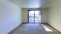 This single-floor residential unit offers a straightforward, functional layout designed for comfortable everyday living. The main living area is finished with wall-to-wall carpeting and neutral-toned walls, creating a clean and versatile space ready for furnishing. A large sliding glass door dominates one wall of the living room, flooding the interior with natural light and providing direct access to a private outdoor deck — an ideal spot for relaxing or enjoying the surrounding views of the neighboring brick building and open sky.

Transitioning toward the back of the unit, the open layout connects the living area to the entryway and kitchen zone. The entry features dark-toned wood doors, adding a warm contrast to the light-colored walls. The kitchen is finished with tile flooring and is equipped with a refrigerator, cabinetry, and an overhead ceiling fan with lighting, providing functional illumination for both the kitchen and adjacent dining or living space.

Throughout the unit, textured ceilings and dark wood trim accents on the doors and window frames add subtle character. The overall ambiance is bright, neutral, and move-in ready, offering a practical living environment with cohesive finishes throughout its single-level floor plan.