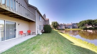 Located at Crooked Creek, this single-floor unit is part of a well-established multi-building residential community characterized by classic neutral-toned siding, pitched rooflines, and decorative chimneys. The exterior presents a tidy, organized aesthetic consistent throughout the neighborhood.

A notable feature of this unit is its ground-level concrete patio, accessed via a sliding glass door, providing a functional outdoor living area ideal for relaxation. The patio sits beneath an elevated deck belonging to the floor above, offering a degree of overhead coverage. The surrounding lawn is well-maintained and generously sized, extending toward a scenic pond that runs along the back of the property. This water feature adds a tranquil natural element to the outdoor environment, enhancing the overall ambiance with reflective views and a sense of openness.

The community setting features mature trees and landscaping throughout, contributing to a established, park-like atmosphere. Adjacent buildings mirror the same architectural style, reinforcing a cohesive neighborhood character. The backyard orientation and pond proximity make this unit particularly well-suited for those who value outdoor scenery and a peaceful residential environment.