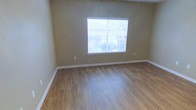 This single-story space presents a clean, straightforward layout designed for comfortable everyday living. The room visible features warm-toned hardwood-style flooring that runs throughout, lending a cohesive and inviting feel underfoot. The walls are finished in a neutral beige tone, providing a versatile backdrop that complements a wide range of furnishing styles and décor choices. A centrally positioned window fitted with horizontal blinds allows natural light to filter into the space, creating a bright and airy atmosphere while offering a view of the outdoor surroundings. White baseboards frame the perimeter of the room, adding a crisp contrast to the warm flooring and walls. The space appears well-maintained and move-in ready, with no visible wear or damage. The overall ambiance is understated and neutral, making it highly adaptable to personal customization. The single-floor layout ensures easy accessibility throughout, with no stairs to navigate. This space would suit those seeking a low-maintenance, functional environment with a clean aesthetic foundation ready to be personalized.
