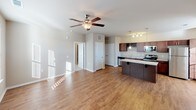 This single-floor unit offers a well-designed open-concept layout that seamlessly connects the kitchen and living areas. The main living space features warm hardwood-style flooring throughout, neutral-toned walls, and a ceiling fan with integrated lighting that enhances both comfort and brightness. Natural light filters in through windows fitted with blinds, contributing to an airy, welcoming ambiance.

The kitchen is a functional centerpiece of the unit, outfitted with rich dark wood cabinetry that includes a distinctive built-in wine rack. Granite-style countertops extend across the workspace and continue onto a peninsula island with elegant pendant lighting overhead. Stainless steel appliances include a gas range, dishwasher, microwave, and refrigerator. Track lighting above the main counter provides focused task illumination.

The kitchen opens directly to the spacious living area, making the layout well-suited for everyday living and entertaining. Additional doors lead to separate rooms, offering defined private spaces within the open floor plan.

The bathroom is equipped with a tub and shower combination, a toilet, and a vanity with a single sink set into a dark granite-style countertop, complemented by a large wall mirror and chrome fixtures. The textured walls and neutral palette carry the cohesive design aesthetic throughout.