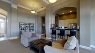 Located at 4610 Weatherford Ln in Columbus, Ohio, this single-floor home showcases a thoughtfully designed open-concept layout that seamlessly connects the living and kitchen areas. The living room is anchored by a coffered ceiling with warm pendant lighting, lending an air of refined elegance to the space. Neutral-toned walls and plush carpeting create a welcoming ambiance, while decorative columns with arched transitions define the boundary between the living and kitchen zones without interrupting the open flow. A built-in shelving unit displays curated décor, adding character and warmth to the room. The kitchen features rich dark countertops set against cream cabinetry and is equipped with stainless steel appliances. A raised bar counter with sleek black barstools provides a convenient casual dining option and serves as a natural gathering point. Recessed lighting throughout the kitchen highlights the deep, contrasting accent wall, creating a cozy and intimate atmosphere. The overall design balances practicality with aesthetic appeal, combining classic architectural details such as crown molding and arched doorways with contemporary finishes for a timeless and inviting home environment.
