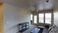 This single-floor residence offers a comfortable and functional layout filled with natural light. The living area serves as the heart of the space, featuring soft carpet flooring and neutral-toned walls that create a clean, understated aesthetic. A ceiling fan with integrated lighting is centrally mounted, providing both air circulation and ambient light to the room.

Large windows flank a glass-paneled exterior door, flooding the living space with abundant natural light and offering views to an outdoor balcony. The balcony access adds an element of openness, extending the livable feel of the interior. The windows are dressed simply, allowing maximum light penetration throughout the day.

The living area provides ample room for seating and furnishings, as evidenced by the open floor plan that comfortably accommodates a sofa, coffee table, and storage shelving. A slim metal shelving unit along one wall offers practical storage without overwhelming the space.

An adjacent room, visible through an interior doorway, suggests additional living or working space connected seamlessly to the main area. The overall ambiance is bright, practical, and move-in ready, making it well-suited for everyday residential use.