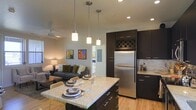 This single-floor space showcases a thoughtfully designed open-concept layout that seamlessly connects the kitchen and living areas. The kitchen is a standout feature, boasting rich dark cabinetry with sleek silver hardware, a stainless steel refrigerator, and a marble-style tile backsplash. A large granite-topped island with seating serves as both a functional prep area and an informal dining space, illuminated overhead by a trio of modern pendant lights. A built-in wine rack adds a refined touch to the upper cabinetry. Light hardwood flooring runs throughout, enhancing the warm, contemporary feel of the space. Recessed lighting and a ceiling fan provide practical comfort across the open floor plan. The living area flows naturally from the kitchen, furnished with a comfortable sofa accented with colorful throw pillows and complemented by warm ambient lighting from a classic table lamp. Colorful wall art adds personality and visual interest to the neutral-toned walls. A glimpse into an adjoining room suggests additional private living space beyond. Overall, the space strikes a balance between modern design and comfortable, everyday livability.