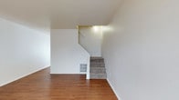 This two-floor space presents a straightforward and functional layout, characterized by its clean lines and neutral palette. The main level showcases warm-toned hardwood-style flooring that extends throughout the open area, creating a welcoming and cohesive foundation. Crisp white walls reflect light effectively, contributing to a bright and airy atmosphere across the lower level.

A carpeted staircase connects the two floors, featuring a simple white railing and balustrade that complement the overall minimalist design. The stairwell is illuminated by a ceiling-mounted light fixture, ensuring the transition between floors remains well-lit. A floor-level vent is visibly integrated into the base of the staircase wall, indicating a built-in HVAC system for climate control throughout the space.

The overall design aesthetic is clean and understated, with an emphasis on practical use of space. The combination of hard flooring on the main level and carpet on the stairs adds both visual contrast and functional comfort. The uncluttered, open feel of the lower level suggests versatile use potential.