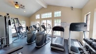 This single-story fitness center offers a bright and functional workout environment designed to accommodate a variety of exercise routines. The space features vaulted ceilings with exposed beams, adding a sense of openness and architectural character. A bank of large, multi-pane windows lines the walls, flooding the interior with natural light while providing views of the surrounding trees and landscaping, creating an inviting atmosphere for exercise.

The facility is equipped with a solid selection of cardio machines, including treadmills from True Fitness, elliptical trainers, and stair climbers, all arranged to maximize floor space and ease of movement. Additional strength and conditioning equipment is visible toward the rear of the room, offering users a well-rounded workout experience. The flooring is a dark, durable material suited for high-traffic fitness use.

The warm beige walls and neutral tones contribute to a clean, focused environment. Security is maintained via a visible camera, and posted signage indicates that facility access requires a key, suggesting this is an amenity space within a managed community. Overall, the fitness center balances functionality with a welcoming, naturally lit design.