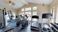This single-story fitness center offers a bright and functional workout environment designed to accommodate a variety of exercise routines. The space features vaulted ceilings with exposed beams, adding a sense of openness and architectural character. A bank of large, multi-pane windows lines the walls, flooding the interior with natural light while providing views of the surrounding trees and landscaping, creating an inviting atmosphere for exercise.

The facility is equipped with a solid selection of cardio machines, including treadmills from True Fitness, elliptical trainers, and stair climbers, all arranged to maximize floor space and ease of movement. Additional strength and conditioning equipment is visible toward the rear of the room, offering users a well-rounded workout experience. The flooring is a dark, durable material suited for high-traffic fitness use.

The warm beige walls and neutral tones contribute to a clean, focused environment. Security is maintained via a visible camera, and posted signage indicates that facility access requires a key, suggesting this is an amenity space within a managed community. Overall, the fitness center balances functionality with a welcoming, naturally lit design.