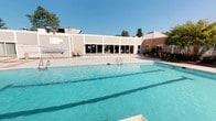This single-story facility is centered around a generously sized outdoor swimming pool, which serves as the defining feature of the property. The pool is filled with clear, blue water and is accessed via stainless steel entry rails, making it both functional and inviting. The surrounding concrete deck provides ample space for lounging and poolside activities, while low-profile pool equipment is visible along the perimeter.

The building adjacent to the pool features a long expanse of floor-to-ceiling sliding glass doors, which create a seamless visual and physical connection between the indoor spaces and the outdoor pool area. This design allows for abundant natural light to fill the interior while maintaining easy access to the deck. The exterior of the building is clad in light-colored siding, contributing to a clean and practical aesthetic. A secondary structure with a brick facade and a metal roof is also visible, suggesting additional functional spaces within the complex.

Mature trees and landscaping with colorful plantings frame the perimeter of the property, adding a touch of greenery and visual warmth to the overall environment. Under clear skies, the outdoor area feels open and bright, making it well-suited for recreational use. The overall design of the facility reflects a practical, community-oriented layout optimized for active outdoor and indoor use.