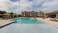 Located at 50 Fenway Place in Concord, North Carolina, this outdoor amenity space is designed to deliver a resort-style living experience. The centerpiece of the area is a large, sparkling swimming pool with crystal-clear blue water, accessible via stainless steel entry rails and featuring a shallow wading area. The pool is flanked by a spacious, textured concrete sun deck that provides ample room for relaxation and social gatherings. Rows of sleek, modern chaise lounge chairs are arranged on artificial turf patches, offering a comfortable spot to unwind under market umbrellas that provide shade throughout the day. A vibrant mural adds an artistic focal point along the pool perimeter. An adjacent clubhouse-style structure with covered outdoor seating, stone accents, and a standing-seam metal roof provides a shaded retreat and appears to connect to interior amenity spaces. The surrounding landscape includes manicured green lawns, ornamental fencing, and mature trees that frame the outdoor environment. Detached garage structures are also visible on the property, contributing to the well-organized layout. The multi-story residential buildings overlooking the pool feature balconies and a craftsman-inspired architectural style, completing the cohesive community aesthetic.