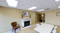 This single-floor space is thoughtfully designed to balance comfort and functionality. The main common area serves as the focal point of the space, featuring a classic white decorative fireplace mantel accented with warm wood trim, topped with framed artwork and decorative vases with dried floral arrangements. A cushioned armchair nearby adds a cozy, inviting touch to this seating area. The room transitions seamlessly into a dining or meeting area, furnished with clean white tables and chairs that provide ample space for group gatherings or activities. The flooring features a light-toned vinyl tile with subtle patterning, contributing to a bright and easy-to-maintain environment. Overhead, a suspended ceiling with recessed panel lighting ensures consistent, even illumination throughout the space. The warm yellow-toned walls create a calm and welcoming atmosphere. Toward the rear of the space, a corridor leads to additional rooms, including windowed interior offices or private rooms accessible through wood-framed doors with sidelights, suggesting a well-organized layout designed to accommodate a variety of uses within the facility.