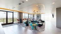 Located at 701 Richmond Ave in Houston's vibrant Montrose neighborhood, this elevated amenity lounge delivers a sophisticated social atmosphere with sweeping panoramic views of the Houston skyline. The open-plan space is defined by a striking curved tray ceiling trimmed in warm gold, fitted with layered ambient lighting and oversized circular pendant fixtures that anchor the room's contemporary aesthetic.

Floor-to-ceiling windows and glass doors flood the interior with abundant natural light while framing expansive outdoor views and providing direct access to a balcony. Large-format porcelain tile flooring transitions into a patterned area rug, delineating a generously sized lounge zone furnished with bold, sculptural seating — including tufted sectional sofas in teal, curved accent chairs in multicolor stripe, and organic-form ottomans that reflect a playful, designer sensibility.

A dedicated kitchenette bar area features a dramatic dark stone island with a fluted wood base, sleek cabinetry in white textured tile, a glass-door beverage refrigerator, and striking wire-cage pendant lights overhead. A bar-height seating area positioned along the window wall offers front-row views of the cityscape. Wall-mounted art and lush tropical plants complete the vibrant, resort-inspired ambiance of this well-appointed communal gathering space.