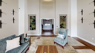 This single-floor amenity space is thoughtfully designed to offer a variety of social and functional areas within a cohesive, contemporary aesthetic. The grand lobby serves as an impressive entry point, featuring soaring double-height ceilings, large floor-to-ceiling glass doors that invite abundant natural light, and striking built-in vertical botanical art panels flanking the main corridor entrance. Warm hardwood-style flooring flows consistently throughout, unifying the space.

The kitchen and bar area is a central highlight, outfitted with expansive granite countertops, dark espresso cabinetry, a stainless steel dishwasher, an undermount sink, and a row of bold matte black pendant lights overhead. Bar-height seating lines the granite island, creating an inviting gathering spot. Multiple wall-mounted televisions enhance the entertainment-friendly atmosphere.

Adjacent lounge areas feature distinct personalities: one showcases a textured mosaic accent wall paired with a sectional sofa, while another offers a built-in white bookcase, a stone-clad fireplace wall, and corridor access to additional rooms. A dining nook with a round table and glass French doors opens directly to an outdoor balcony with open views. Throughout, warm neutral tones, curated wall art, and recessed lighting contribute to a polished, welcoming ambiance.