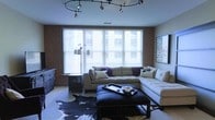This thoughtfully designed single-floor space blends urban industrial character with contemporary comfort. The living area serves as the centerpiece, anchored by exposed red brick walls that add texture and warmth to the room. A bold circular track lighting fixture overhead provides both functional illumination and a striking design statement, casting a flattering glow across the space. Large windows dressed with horizontal blinds span a generous portion of the wall, allowing ample natural light to filter in while offering privacy and light control. The living area is furnished with a plush, tufted sectional sofa in neutral tones, accented with decorative pillows, and complemented by a sleek black ottoman coffee table with nailhead trim that doubles as a stylish centerpiece. A distinctive cowhide area rug grounds the seating arrangement, adding a touch of eclectic flair. A floor lamp with an adjustable arm provides additional ambient lighting, while a low-profile dresser and a framed piece of wall art contribute to the room's polished, curated aesthetic. The overall ambiance is inviting and modern, striking a confident balance between industrial edge and residential comfort.