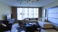 This thoughtfully designed single-floor space blends urban industrial character with contemporary comfort. The living area serves as the centerpiece, anchored by exposed red brick walls that add texture and warmth to the room. A bold circular track lighting fixture overhead provides both functional illumination and a striking design statement, casting a flattering glow across the space. Large windows dressed with horizontal blinds span a generous portion of the wall, allowing ample natural light to filter in while offering privacy and light control. The living area is furnished with a plush, tufted sectional sofa in neutral tones, accented with decorative pillows, and complemented by a sleek black ottoman coffee table with nailhead trim that doubles as a stylish centerpiece. A distinctive cowhide area rug grounds the seating arrangement, adding a touch of eclectic flair. A floor lamp with an adjustable arm provides additional ambient lighting, while a low-profile dresser and a framed piece of wall art contribute to the room's polished, curated aesthetic. The overall ambiance is inviting and modern, striking a confident balance between industrial edge and residential comfort.