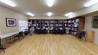 The craft room is a generously proportioned, single-level space designed to support a wide range of creative activities. The room's most prominent feature is the expansive wall of dark wood built-in shelving that lines the perimeter, offering numerous open cubbies filled with labeled storage bins, totes, and craft supplies organized for easy access. Work surfaces are arranged throughout the room in the form of long rectangular tables flanked by black chairs, accommodating multiple participants simultaneously for group crafting sessions.

The flooring throughout is light-toned hardwood-style planking, lending a clean and bright aesthetic to the space. Overhead, flush-mounted fluorescent light fixtures provide consistent, even illumination across the entire room. Natural light is introduced through several windows fitted with horizontal blinds, allowing for adjustable brightness. Framed artwork adorns select walls, adding a decorative touch to the functional environment.

A dedicated corner area includes upper cabinetry, a small desk, and additional storage, offering a more private workspace. A door with an interior window provides access to an adjacent area, while HVAC vents and a tower fan ensure comfortable climate control. The room's open central floor space allows for flexible arrangement to suit varying group sizes and project needs.