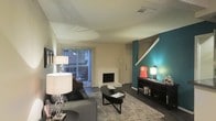This two-story residence in Bedford, Texas offers a thoughtfully designed living space with distinct character. The main living area serves as the focal point of the home, showcasing a combination of neutral tones accented by a bold teal feature wall that adds a vibrant pop of color to the space. A fireplace anchors one side of the room, providing both a functional and visual centerpiece. Sliding glass doors span a significant portion of the exterior wall, allowing generous natural light to filter in while offering a view and direct access to the outdoor area. The living room accommodates comfortable furnishings arranged around a central area rug, creating a welcoming and functional layout. A staircase is visibly integrated into the living area, connecting the main floor to the upper level and contributing to the open, vertical sense of space. Dark hardwood-style flooring grounds the room and contrasts effectively with the lighter walls and ceiling. The textured ceiling adds a subtle dimensional quality to the interior. Overall, the space balances everyday functionality with distinctive design touches throughout.