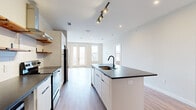 This thoughtfully designed single-floor unit showcases a clean, contemporary aesthetic throughout, unified by warm-toned hardwood-style flooring and crisp white walls. The heart of the space is an open-concept kitchen and living area, where a generous central island with a dark stone countertop and matte black sink serves as a natural gathering point. The kitchen is equipped with stainless steel appliances, including a refrigerator and range, complemented by a stainless hood vent, white cabinetry, open wood shelving, and a classic white tile backsplash. Track lighting overhead highlights the kitchen's modern finishes. French doors at the far end of the living area invite natural light deep into the space, creating an airy and welcoming ambiance. The bedroom areas are well-proportioned, featuring large windows that provide additional natural light. A dedicated bathroom offers a dual-sink vanity with dark countertops, white cabinetry, matte black hardware, and framed mirrors with coordinating wall-mounted lighting — a cohesive and stylish design. A separate, dedicated laundry room houses a full-size washer and dryer, adding a practical and convenient amenity. The unit's consistent use of modern finishes, neutral tones, and purposeful layout make it a highly functional and visually appealing space throughout.