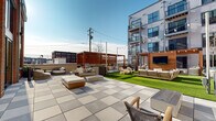 Located at 600 West State Street in Kennett Square, Pennsylvania, this thoughtfully designed outdoor amenity courtyard offers a versatile gathering space surrounded by a striking mixed-material building façade featuring red brick, light masonry, and large-format windows. The space is organized into several distinct zones, each serving a different social function while maintaining a cohesive contemporary aesthetic.

Large-format paver tiles define the primary hardscape areas, creating clean, durable walkways and lounging platforms throughout. A lush artificial turf lawn anchors the central zone, providing a bright, low-maintenance green space ideal for recreation and relaxation. A dedicated ping-pong table sits along one side, adding a recreational focal point.

A warm wood pergola structure serves as a centerpiece, housing an outdoor living room arrangement complete with plush sectional seating and a wall-mounted television — ideal for communal viewing and entertaining. Nearby, a covered outdoor kitchen and bar area with high-top seating offers a functional space for cooking and casual dining.

String lights crisscross overhead throughout the courtyard, lending an inviting ambiance during evening hours. Multiple seating vignettes with modern wood-framed furniture and neutral cushions are distributed across the space, providing ample options for both small gatherings and larger group events. The open-sky setting ensures abundant natural light throughout the day.