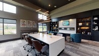 Located at 9295 One Deerfield Place in Mason, Ohio, this single-floor amenity lounge delivers a sophisticated blend of collaborative workspace and comfortable social seating. The open-plan layout is anchored by a large white central conference table surrounded by tan leather task chairs, ideal for meetings or focused work. Dramatic linear pendant lighting suspended from the dark ceiling adds a striking visual element above the workspace.

Along one wall, a built-in dark wood shelving unit frames a sleek linear fireplace clad in textured stone, with a mounted display above. This area transitions into a relaxed lounge zone furnished with plush teal barrel chairs and natural wood stump accent tables, creating an inviting space for informal gatherings. A dedicated alcove features a tufted upholstered bench with sconce lighting flanked by individual desks, offering semi-private nooks for independent work.

The space benefits from an elevated ceiling height with clerestory windows along the upper walls, allowing natural light to fill the interior while maintaining privacy. Colorful abstract artwork adorns the walls, reinforcing the space's contemporary aesthetic. Patterned area rugs help define each zone while contributing texture and warmth throughout this thoughtfully designed, multifunctional environment.