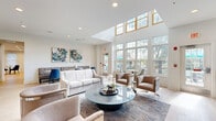 Located at 17W720 Butterfield Rd in Oakbrook Terrace, IL, this two-floor amenity facility offers a thoughtfully designed and well-appointed space for residents and guests alike. The ground floor welcomes visitors through a dedicated entrance that opens into bright, open common areas finished with light hardwood-style flooring and crisp white walls. The main lounge is an architectural highlight, boasting soaring double-height ceilings, a dramatic skylight, and walls of large windows that flood the space with natural light and frame views of the surrounding greenery. Comfortable seating arrangements, a glass-top coffee table, and vibrant artwork create an inviting social atmosphere. Adjacent dining and lounge nooks feature stylish banquette seating, including a bold accent wall booth and casual round tables with modern blue chairs. The kitchen is equipped with sleek dark cabinetry, white tile backsplash, and premium under-counter refrigeration. A fully equipped fitness center offers cardio and strength training equipment, wall-to-wall mirrors, and abundant natural light. A dedicated yoga and movement studio features light hardwood floors, full-length mirrors, and ceiling fans. The upper floor hosts additional multipurpose rooms, including glass-walled private office or meeting spaces with carpet tile flooring, accessible via staircase or elevator. Throughout, the design balances modern finishes with a clean, airy aesthetic.