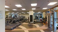 This modern fitness center is designed to accommodate a comprehensive workout experience within a single, open-plan floor. The space is outfitted with a broad selection of cardio equipment, including treadmills and elliptical machines, arranged in neat rows along one side of the room. A dedicated free weights area features a full rack of dumbbells and medicine balls of varying sizes, catering to both strength and functional training needs. At the center of the room, an adjustable workout bench adds versatility for resistance exercises. Large, floor-to-ceiling mirrors line the walls, visually expanding the space and allowing exercisers to monitor their form throughout their sessions. The flooring features a bold, curvilinear design in warm earth tones, adding a dynamic and energetic character to the environment. Overhead, a grid of recessed panel lighting ensures the entire space is evenly and brightly illuminated, creating a clean and motivating atmosphere. Natural light filters in through windows along one wall, further enhancing the open and airy feel of the facility. The overall layout is thoughtfully organized to maximize usability while maintaining comfortable circulation throughout the space.