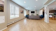 Located at 1070 Noëlla Leclair Way in Ottawa, Ontario, this single-floor community space offers a thoughtfully designed layout that balances comfort, function, and social interaction. Light wood-look flooring flows seamlessly throughout, creating a warm and cohesive aesthetic across all areas.

The main living room is a generously sized, open area bathed in natural light from multiple windows fitted with horizontal blinds. A wall-mounted flat-screen television anchors the seating arrangement, which includes a large grey sectional sofa and coordinating blue accent chairs, centered around an electric fireplace. Recessed ceiling lighting and a wall-mounted air conditioning unit add to the room's practicality and comfort.

An adjacent casual lounge area features round white tables with modern Eames-style chairs and decorative green plants, lending a fresh and inviting atmosphere. A classic arcade cabinet adds a playful and entertaining element to the space.

The dedicated office is a compact, private room accessed through a glass-panel door with a sleek metal handle. It is furnished with a wooden desk and contemporary chrome-framed chairs, making it well-suited for focused work or private meetings.

The entry area is clean and functional, with recessed lighting, a secured door, and nature-inspired wall art that sets a welcoming tone. Throughout the space, Canadian-themed photography adds a distinctive local character.