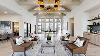 This amenity center is designed to offer a comprehensive range of social and recreational spaces within a single, thoughtfully arranged floor. The centerpiece is a grand clubhouse lounge distinguished by soaring vaulted ceilings with exposed beam detailing, a dramatic multi-shade pendant chandelier, and expansive black-framed windows that flood the interior with natural light and provide views of the outdoor pool area. Rich brown leather seating arrangements and layered area rugs anchor the space, while a built-in wet bar with wood cabinetry, dark tile backsplash, open shelving, and stainless steel appliances provides a convenient refreshment station nearby. A secondary kitchenette features granite countertops, hexagonal tile flooring, and pendant lighting for additional functionality. The fitness center is generously proportioned, equipped with cardio machines, strength equipment, and a dedicated stretching area with stability balls and a wall-mounted TV. A comfortable media lounge offers plush sectional seating on patterned carpeting with a large flat-screen display. A private conference room with a formal table and globe pendant lighting accommodates meetings. Outdoors, a resort-style pool with in-water lounge chairs is complemented by a pergola-covered seating area surrounded by manicured landscaping.