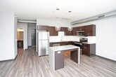 This single-floor unit delivers a well-appointed, modern living experience with a thoughtful open-concept layout. The kitchen serves as a central highlight, showcasing rich dark-wood cabinetry, light granite-style countertops, a classic white subway tile backsplash, and a full suite of stainless steel appliances including a refrigerator, range, microwave, and dishwasher. A generously sized island with pendant globe lighting above anchors the kitchen and provides additional prep and seating space. Exposed ductwork along the ceiling lends an industrial-loft character to the open living and dining area, which flows seamlessly from the kitchen. Wood-look plank flooring runs throughout the main living spaces, adding warmth and visual continuity. The bedroom features plush carpet, neutral gray walls, and a dedicated walk-in closet with built-in shelving and hanging rods, offering ample storage. The bathroom is clean and functional, featuring a tiled tub-shower combination with a curved curtain rod, a dark-vanity with a granite-style countertop, and brushed nickel fixtures. Throughout the space, the design palette—neutral tones, dark accents, and modern finishes—creates a cohesive, contemporary aesthetic.