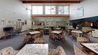 This welcoming community activity room is defined by its dramatic double-height ceilings accented with warm wood beams, creating an open and airy atmosphere. A generous band of clerestory windows runs along the upper walls, flooding the space with natural light and offering views of surrounding trees, enhancing the connection to the outdoors. Ceiling fans are positioned throughout to ensure year-round comfort.

The main gathering area is thoughtfully furnished with numerous square tables and floral-upholstered swivel chairs on casters, arranged to encourage both small group interaction and larger social gatherings. A jigsaw puzzle in progress on one of the tables speaks to the space's leisurely, community-focused purpose. Artwork adorns the walls throughout, adding color and personality to the neutral palette of whites and grays.

Toward the back of the room, a pool or billiards table provides additional recreational options, while a dedicated area with shelving and a display screen serves as a resource or media corner. A teal accent wall adds a pop of color, visually anchoring a cozy reading or library nook with bookshelves. The neutral carpeted flooring ties the entire space together, making this a versatile and inviting hub for community life.