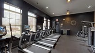 This fitness centre in Brighton 3050 is a comprehensively equipped single-level facility designed to support a full range of training goals. The space is divided into distinct functional zones, each thoughtfully arranged to maximise usability and flow.

The cardio zone features a row of True-brand treadmills and elliptical machines, each equipped with individual digital consoles. A wall-mounted flat-screen television overlooks this area, and multiple windows fitted with blinds allow natural light to filter in while maintaining a focused atmosphere. Dark charcoal-painted walls and recessed ceiling lighting contribute to a modern, high-contrast aesthetic throughout this section.

The strength training area transitions into a warmer, neutral-toned environment, where a full rack of dumbbells is displayed in front of a large wall mirror, enhancing the sense of space. Stability balls and benches provide versatility for free weight exercises. Adjacent to this, a dedicated machine training zone houses cable and pulley systems along with selectorised strength equipment.

Rubber flooring throughout ensures durability and safety underfoot. A beverage refrigerator, wall clock, and pendant lighting add practical and decorative touches. Windows overlooking adjacent rooms provide visual connectivity within the broader facility.