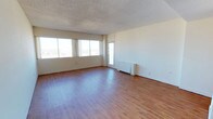 This single-floor space offers an open, versatile layout highlighted by warm hardwood-style flooring that extends throughout the main area. Large windows along one wall flood the interior with abundant natural light, providing views of the surrounding exterior while creating a bright and airy atmosphere. A glass-paneled door adds an additional source of light and offers direct access to the outdoors. The walls are finished in a clean, neutral white, providing a blank canvas adaptable to a wide range of purposes. A textured popcorn ceiling adds a classic architectural detail overhead. A baseboard heating unit positioned along the far wall ensures functional climate control. The open floor plan allows for flexible arrangement, with generous square footage that comfortably accommodates multiple configurations. The overall ambiance is clean, well-lit, and straightforward, making this space well-suited for residential or light commercial use.