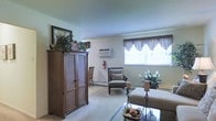 This single-floor space offers a comfortable and functional living area finished in soft, neutral tones throughout. The living room is carpeted in a light beige, contributing to a warm and inviting ambiance. Natural light filters in through a large window dressed with decorative valance curtains, brightening the room without overwhelming it. A wall-mounted air conditioning unit provides climate control, complemented by a baseboard heating system along the perimeter walls.

The room is well-suited for relaxed living, with space enough to accommodate a substantial wooden armoire with woven panel doors, an upholstered accent chair, and a glass-topped coffee table. Decorative touches, including potted greenery and a bowl of decorative accents on the coffee table, add character to the space. The smooth, light-colored walls and standard ceiling height maintain an open, uncluttered feel. Overall, the space presents a traditionally styled, practical layout ideal for everyday living, offering a balance of functional storage, comfortable seating, and understated decorative appeal.