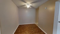 This single-story space presents a clean, functional interior centered around a well-proportioned room visible in the image. The flooring consists of rich, dark hardwood-style planks that add warmth and contrast to the neutral gray and beige-toned walls. White baseboards run along the perimeter, providing a crisp boundary between the walls and floor. The ceiling features a textured finish and is equipped with a white ceiling fan with an integrated light fixture, offering both ventilation and overhead illumination. A doorway is visible along one wall, suggesting a straightforward and practical layout. A smoke or carbon monoxide detector is mounted on the wall, noting attention to safety features. The overall ambiance is simple, neutral, and move-in ready, with a palette that provides a versatile backdrop suitable for a variety of furnishing styles. The space conveys a modest yet comfortable atmosphere, ideal for everyday living.