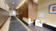 This well-appointed lobby space makes a strong architectural statement through its standout geometric wood panel feature wall, where overlapping rectangular forms in warm brown tones create a layered, three-dimensional effect that immediately draws the eye. The design is both modern and inviting, combining clean lines with rich material textures. Ambient recessed lighting and a sleek linear ceiling light strip illuminate the corridor, casting a warm glow across the space and highlighting the craftsmanship of the feature wall. The flooring transitions from a textured tile plank base along the walls to a patterned carpet runner along the main corridor, lending both visual interest and functional durability to the high-traffic area. A white upholstered armchair is positioned near the feature wall, offering a comfortable waiting area for visitors. A bank of individual mailboxes is mounted on the adjacent wall, indicating this lobby serves a multi-unit residential or mixed-use building. At the far end of the corridor, a stainless steel elevator with clearly marked signage provides vertical access throughout the building. The overall ambiance is professional yet welcoming, with a cohesive color palette of warm yellows, browns, and neutral grays contributing to a refined and contemporary aesthetic.