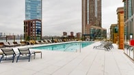Located in Long Island City, Queens, this rooftop amenity deck at 2-20 Malt Dr offers an impressive outdoor retreat above the urban landscape. The centerpiece is a generously sized rectangular swimming pool with crystal-clear turquoise water, framed by warm cedar and glass architectural elements that lend a refined, resort-like character to the space.

Surrounding the pool is a broad, light-tiled sundeck lined with rows of contemporary lounge chairs and side tables, providing ample space for relaxation. A dedicated pergola structure with a louvered roof sits adjacent to raised planter beds filled with lush seasonal greenery, offering a shaded seating area with Adirondack-style chairs for a more intimate gathering spot.

An outdoor communal dining area features long wooden picnic tables with bench seating, framed by ornamental grasses and verdant planters — ideal for social gatherings and al fresco dining.

The deck's perimeter is enclosed by a glass-paneled railing system, ensuring unobstructed panoramic views of the Manhattan skyline, the East River, and the surrounding Long Island City high-rise developments. The overall ambiance is that of a polished, urban rooftop escape, combining functional amenities with thoughtfully designed landscaping and striking cityscape vistas.