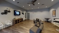 This single-floor communal space is thoughtfully designed to serve as a social hub, blending functional areas with comfortable, inviting aesthetics throughout. The layout flows seamlessly across several distinct zones, all unified by warm wood-look flooring and neutral wall tones.

The lounge and media area anchors one end of the space, featuring a wall-mounted flat-screen television, recessed lighting, and a ceiling fan overhead. Comfortable modular seating with built-in side tables is arranged for optimal viewing, while additional bistro-height tables and chairs provide flexible gathering options. Classic framed movie posters — including Rocky and Star Wars — line the walls, lending the room a vibrant, entertainment-focused character.

Adjacent to the lounge is a well-appointed kitchenette, designed with practicality in mind. Expansive upper and lower cabinetry in a contemporary wood-grain finish lines the walls in an L-shaped configuration, offering generous storage. Light gray countertops provide ample prep and serving surface, complemented by a stainless steel refrigerator and a built-in sink. Natural light filters in through windows, brightening the space during daytime hours.

French doors with glass pane inserts connect the various zones, allowing light to pass freely and maintaining an open, airy feel throughout. The overall design balances modern finishes with a warm, welcoming ambiance well-suited for community gatherings and social activities.