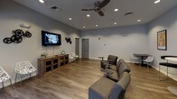 This single-floor communal space is thoughtfully designed to serve as a social hub, blending functional areas with comfortable, inviting aesthetics throughout. The layout flows seamlessly across several distinct zones, all unified by warm wood-look flooring and neutral wall tones.

The lounge and media area anchors one end of the space, featuring a wall-mounted flat-screen television, recessed lighting, and a ceiling fan overhead. Comfortable modular seating with built-in side tables is arranged for optimal viewing, while additional bistro-height tables and chairs provide flexible gathering options. Classic framed movie posters — including Rocky and Star Wars — line the walls, lending the room a vibrant, entertainment-focused character.

Adjacent to the lounge is a well-appointed kitchenette, designed with practicality in mind. Expansive upper and lower cabinetry in a contemporary wood-grain finish lines the walls in an L-shaped configuration, offering generous storage. Light gray countertops provide ample prep and serving surface, complemented by a stainless steel refrigerator and a built-in sink. Natural light filters in through windows, brightening the space during daytime hours.

French doors with glass pane inserts connect the various zones, allowing light to pass freely and maintaining an open, airy feel throughout. The overall design balances modern finishes with a warm, welcoming ambiance well-suited for community gatherings and social activities.