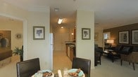 This single-floor residence is designed with an open-concept layout that connects the dining area, kitchen, and living space in a cohesive, functional arrangement. The dining area serves as a central hub, featuring dark-framed chairs and a table set with decorative dishware, positioned to take advantage of sightlines throughout the space. Neutral, warm tones dominate the walls and flooring, contributing to a welcoming ambiance.

The kitchen, visible through a wide corridor, is outfitted with medium-toned wood cabinetry, dark countertops, and standard appliances including a full-size refrigerator. A double sink is positioned along the countertop run, and overhead lighting illuminates the workspace effectively. Crown molding accents the ceiling in the main living areas, adding a refined architectural detail. Framed artwork on the walls provides visual interest without overwhelming the understated decor.

Adjacent to the dining space, the living area accommodates seating and benefits from natural light filtering in through a window. The textured ceiling and light-toned flooring throughout create a consistent, clean aesthetic across all visible areas of the residence.