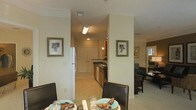 This single-floor residence is designed with an open-concept layout that connects the dining area, kitchen, and living space in a cohesive, functional arrangement. The dining area serves as a central hub, featuring dark-framed chairs and a table set with decorative dishware, positioned to take advantage of sightlines throughout the space. Neutral, warm tones dominate the walls and flooring, contributing to a welcoming ambiance.

The kitchen, visible through a wide corridor, is outfitted with medium-toned wood cabinetry, dark countertops, and standard appliances including a full-size refrigerator. A double sink is positioned along the countertop run, and overhead lighting illuminates the workspace effectively. Crown molding accents the ceiling in the main living areas, adding a refined architectural detail. Framed artwork on the walls provides visual interest without overwhelming the understated decor.

Adjacent to the dining space, the living area accommodates seating and benefits from natural light filtering in through a window. The textured ceiling and light-toned flooring throughout create a consistent, clean aesthetic across all visible areas of the residence.