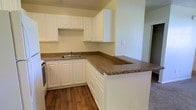 This single-story space showcases a well-appointed kitchen as its central feature. The kitchen is designed with an abundance of white raised-panel cabinetry that extends to the upper walls, providing generous storage throughout. The cabinetry is complemented by rich, granite-style countertops in warm earth tones featuring brown and gold flecks, which extend across the workspace and wrap around a peninsula, creating a natural division between the kitchen and the adjacent area. This peninsula offers additional prep space and could comfortably serve as a casual dining or serving area. The kitchen includes a stainless steel sink positioned along the main counter run, with space accommodating standard appliances. Warm-toned hardwood flooring runs throughout the kitchen, adding a welcoming and cohesive aesthetic to the space. The walls are finished in a soft, neutral yellow-green tone that pairs well with the white cabinetry. A doorway visible in the background hints at additional rooms beyond the kitchen, suggesting a practical and connected floor plan. The overall atmosphere is clean, functional, and move-in ready.