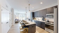This thoughtfully designed single-floor residence combines contemporary style with functional living. The open-concept layout seamlessly connects the kitchen, dining, and living areas, creating a bright and cohesive space bathed in natural light. The kitchen is a standout feature, showcasing dark wood cabinetry paired with a marble-style tile backsplash, stainless steel appliances, and a generous island with a white quartz countertop and seating for multiple guests. Globe pendant lights with brass accents add a refined finishing touch overhead. Light hardwood flooring flows throughout the main living area, complementing the clean white walls and creating a warm, inviting ambiance. The living room opens through floor-to-ceiling sliding glass doors to a private balcony, offering pleasant outdoor views. The bedroom is a calm retreat, featuring a large window with sheer curtains that allow soft natural light, and access to a well-organized walk-in closet with built-in shelving and hanging space. The bathroom is finished with a white vanity, black hardware accents, a large framed mirror, and wood-toned tile surrounds. Every detail of this residence reflects a modern aesthetic with a warm and welcoming character.