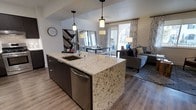 This single-floor space showcases a thoughtfully designed open-concept layout that seamlessly connects the kitchen, dining, and living areas. The kitchen serves as a central hub, anchored by a substantial island topped with light-veined granite countertops and equipped with a undermount sink and stainless steel dishwasher. Dark cabinetry beneath the island provides ample storage while creating a striking contrast against the stone surface. Pendant lighting hangs above the island, adding focused illumination and a contemporary aesthetic detail.

The dining area sits adjacent to the kitchen, benefiting from the same warm pendant fixtures overhead and a decorative mirror that amplifies the sense of depth and light in the space. Hardwood-style flooring flows continuously throughout, unifying the open floor plan and adding warmth underfoot.

The living area is bathed in natural light from large windows dressed with patterned curtains. A comfortable seating arrangement anchors the space, complemented by a textured area rug. The overall ambiance is modern and cohesive, with a neutral color palette, clean lines, and carefully chosen fixtures that create an inviting, well-appointed environment.