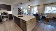 This single-floor space showcases a thoughtfully designed open-concept layout that seamlessly connects the kitchen, dining, and living areas. The kitchen serves as a central hub, anchored by a substantial island topped with light-veined granite countertops and equipped with a undermount sink and stainless steel dishwasher. Dark cabinetry beneath the island provides ample storage while creating a striking contrast against the stone surface. Pendant lighting hangs above the island, adding focused illumination and a contemporary aesthetic detail.

The dining area sits adjacent to the kitchen, benefiting from the same warm pendant fixtures overhead and a decorative mirror that amplifies the sense of depth and light in the space. Hardwood-style flooring flows continuously throughout, unifying the open floor plan and adding warmth underfoot.

The living area is bathed in natural light from large windows dressed with patterned curtains. A comfortable seating arrangement anchors the space, complemented by a textured area rug. The overall ambiance is modern and cohesive, with a neutral color palette, clean lines, and carefully chosen fixtures that create an inviting, well-appointed environment.