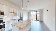 Located at 6300 Clayton Ave in St. Louis, Missouri, this contemporary single-floor residence showcases a thoughtfully designed open-concept layout with high-quality finishes throughout. The kitchen is a standout feature, appointed with crisp white shaker cabinetry, striking granite countertops on both the perimeter surfaces and a generously sized central island, and a full suite of stainless steel appliances including a refrigerator, range, microwave, and dishwasher. Elegant glass globe pendant lights hang above the island, complementing the subway tile backsplash and adding a refined touch to the space. The living area flows seamlessly from the kitchen and is bathed in natural light through expansive floor-to-ceiling sliding glass doors that open directly onto a private balcony, offering scenic treetop and neighborhood views. Wood-style flooring runs throughout the main living spaces, while additional rooms feature light hardwood-style floors and large windows that invite further natural light. The primary bedroom suite connects to a walk-in closet and an en-suite bathroom complete with a tiled walk-in shower and vanity. The overall aesthetic is clean, bright, and modern, delivering a refined residential experience in a well-appointed St. Louis setting.