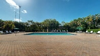 Located at 20891 NW Ninth Ct in Miami Gardens, Florida, this single-story community facility offers a well-appointed outdoor amenity space designed for relaxation and recreation in a lush South Florida setting. The centerpiece of the property is a generously sized rectangular swimming pool, surrounded by a tasteful brick-paver pool deck that provides ample space for lounging. Poolside seating includes multiple chaise lounges and chairs, shaded by umbrellas for comfort in the Florida sun. The pool is equipped with standard safety features including entry handrails and a life ring station. Framing the entire area is a dense canopy of mature tropical palms and lush greenery, creating a private, resort-like atmosphere. A well-maintained lawn area adjoins the pool deck, accented by pathway lighting fixtures that illuminate the grounds after dark. The single-story clubhouse building anchors the space with a warm Mediterranean-inspired architectural style, featuring a distinctive terracotta tile roof, stucco exterior, and an open, covered patio that overlooks the pool. The overall ambiance is tranquil and inviting, blending manicured outdoor spaces with the natural beauty of tropical Florida flora.