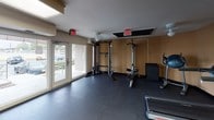 Located at 6503 Landover Rd in Cheverly, Maryland, this single-floor fitness center is designed to provide a well-rounded workout experience. The space is divided into distinct exercise zones, each outfitted with a variety of equipment to accommodate different training needs.

The cardio area features multiple treadmills and elliptical machines arranged in an orderly row, illuminated by flush-mounted ceiling light panels that ensure a bright, energizing environment. Warm-toned walls complement the dark rubber flooring throughout, providing both durability and comfort underfoot.

The strength training area is equipped with a multi-function cable and weight machine, an adjustable bench, and additional resistance equipment. This section benefits from large windows that invite natural light and offer views of the surrounding outdoor greenery, while a ceiling fan promotes air circulation. A secondary strength zone near the entrance provides further equipment options with direct access to the exterior through glass doors.

The facility also includes an accessible restroom with grab bars, tile flooring, and a clean, functional layout designed to accommodate all users.

Overall, the fitness center presents a practical and organized environment with ample equipment variety, good natural lighting, and thoughtful accessibility features, making it a comprehensive amenity for residents of the Cheverly community.