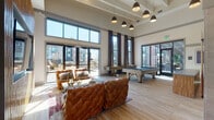 This single-floor communal lounge space is designed with a modern yet warm aesthetic, combining industrial and upscale interior elements throughout. Soaring ceilings with exposed wood beams and large floor-to-ceiling windows flood the interior with natural light, while oversized sliding glass doors connect seamlessly to an outdoor patio area with views of the surrounding neighborhood.

The main gathering area is anchored by a pair of richly tufted cognac leather sofas arranged around a sleek metal-framed coffee table, complemented by a textured bamboo accent wall housing a large flat-screen television. A cluster of stylish pendant lights hangs above, adding warm ambient illumination. Nearby, a full-size pool table and shuffleboard table provide recreational options, making the space equally suited for leisure and entertainment.

A dedicated reception and co-working zone near the entrance features a front desk, workstation computers, and a comfortable seating arrangement, ideal for business or administrative use. Adjacent to this is a well-appointed wet bar kitchenette with marble countertops, wood cabinetry, a gold-accented faucet, built-in microwave drawers, and bar-height seating.

Tucked into an alcove is an intimate lounge nook, defined by dramatic floor-to-ceiling teal velvet upholstered panels, a built-in bench seat, geometric ottomans, and round side tables—offering a quiet retreat within the larger space. Light wood flooring ties all areas together cohesively.